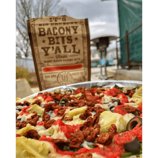 A vibrant vegan pizza topped with artichokes, olives, and plant-based bacon bits from It’s Big Crunchy Bacony Bits Y’all, showcasing a hearty, colorful dish outdoors.
