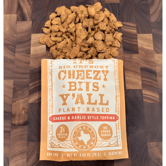 A bag of Big Crunchy Cheezy Bits Y’all with plant-based cheesy and garlic toppings displayed with a generous heap of the product.