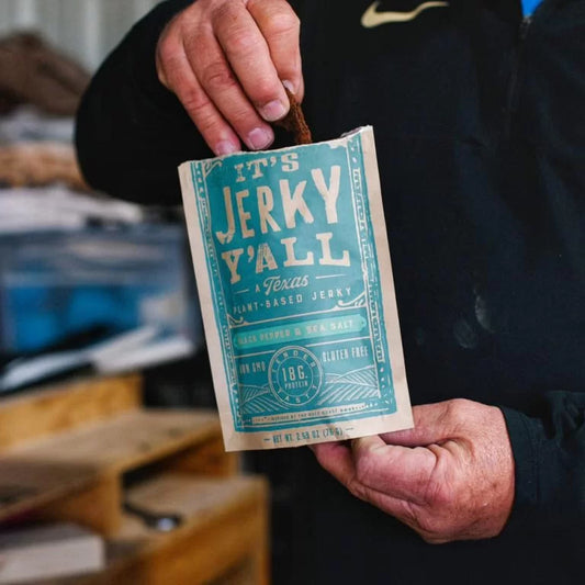A person holding a bag of All Y'all's Foods Black Pepper & Sea Salt plant-based jerky with rustic background elements visible.