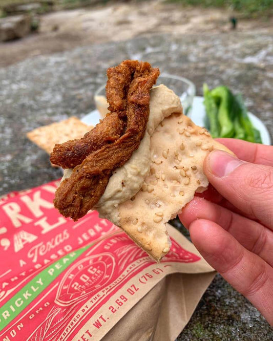 A close-up of a hand holding a sesame cracker topped with hummus and vegan jerky from a bag of 'It's Jerky Y'all' by All Y'all's Foods, highlighting a tasty plant-based snack outdoors.