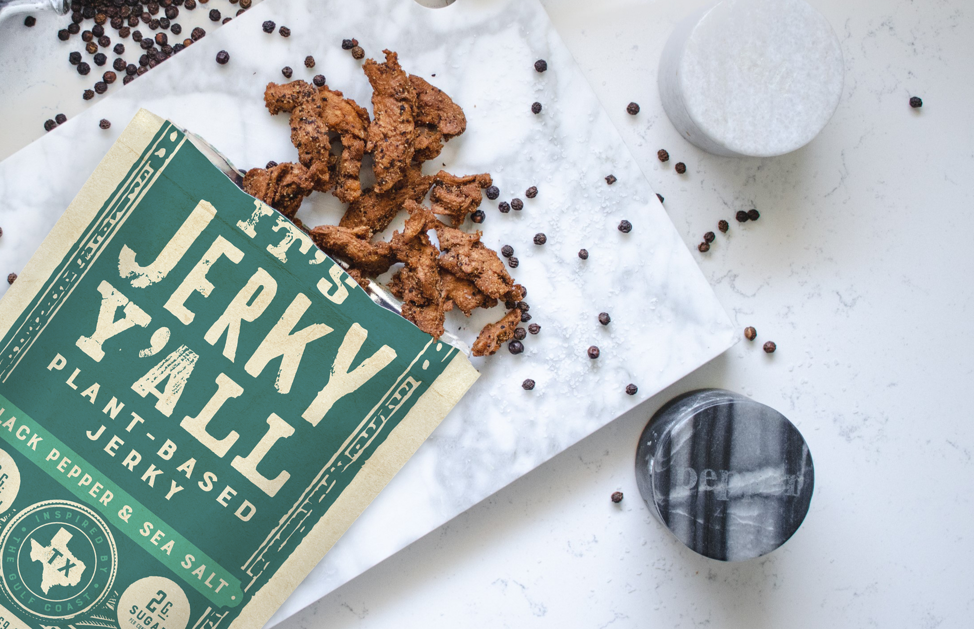 Open bag of 'It's Jerky Y'all' Black Pepper & Sea Salt vegan jerky, a gluten-free vegan snack, displayed on a marble board with black peppercorns.