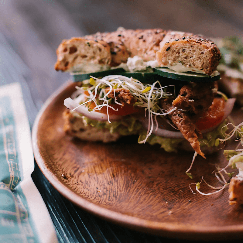 A gourmet bagel sandwich loaded with All Y’alls Foods vegan jerky, fresh vegetables, and sprouts, served on a wooden plate.