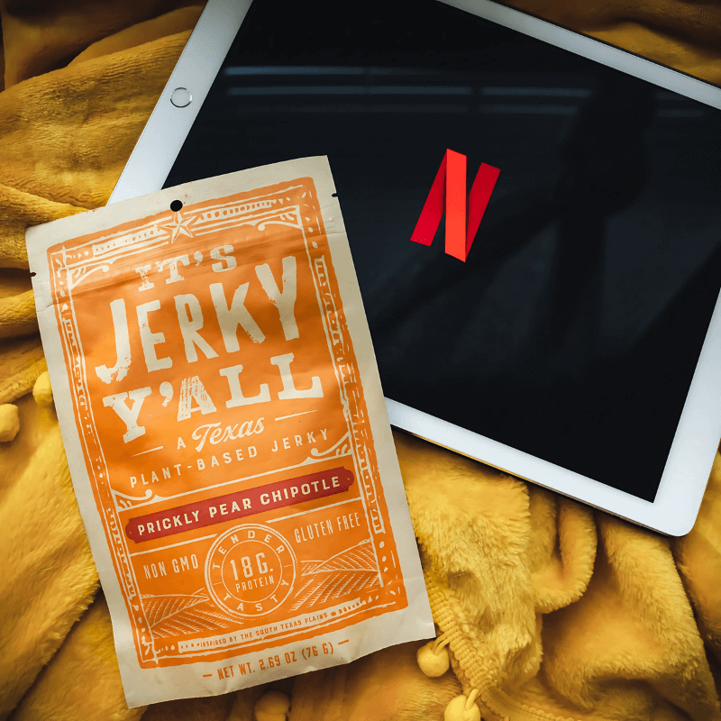 A packet of Prickly Pear Chipotle vegan jerky resting on a cozy yellow blanket beside a tablet displaying the Netflix logo, perfect for a plant-based movie night snack.