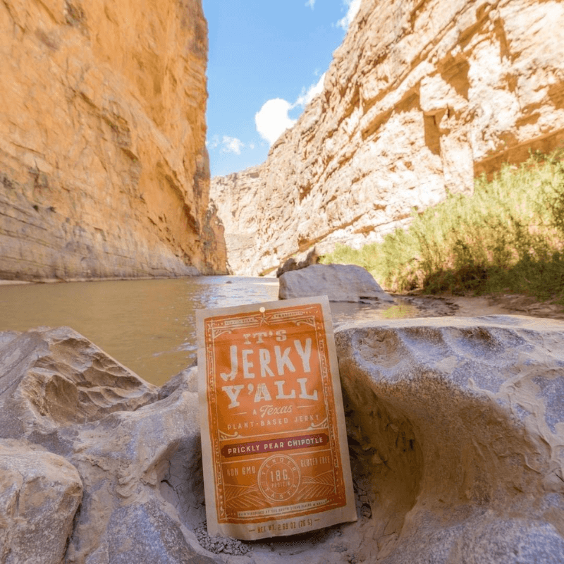 A package of "It's Jerky Y'all" Prickly Pear Chipotle plant-based jerky resting on a rocky ledge in a picturesque desert canyon.