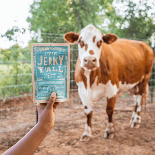 A vibrant image featuring a hand holding a package of "It's Jerky Y'all" Black Pepper & Sea Salt vegan jerky in front of a grazing cow, promoting plant-based alternatives at https://allyallsfoods.com/.