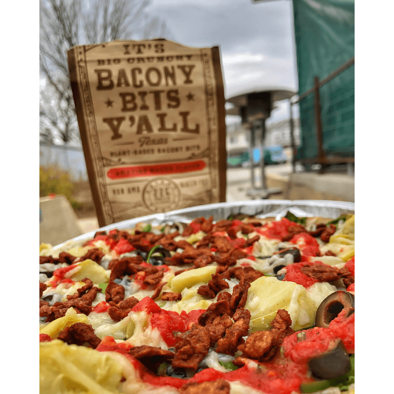 A vibrant vegan pizza topped with artichokes, olives, and plant-based bacon bits from It’s Big Crunchy Bacony Bits Y’all, showcasing a hearty, colorful dish outdoors.