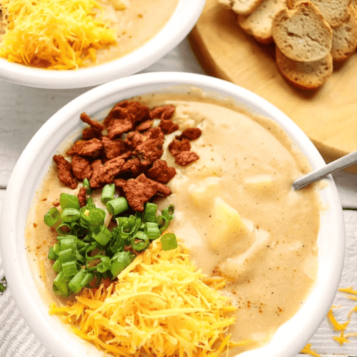 A vibrant bowl of loaded vegan potato soup garnished with green onions, vegan cheese, and All Y’alls Foods' smoky plant-based jerky, showcasing comfort food at https://allyallsfoods.com/.