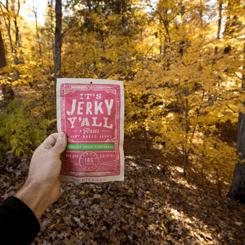 A hand holding a package of plant-based jerky against a vibrant autumnal forest filled with golden leaves.