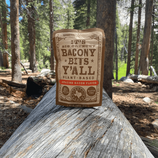 A package of plant-based jerky, "Big Crunchy Bacon Bits Y’all," sitting on a rustic log in a scenic forest setting, highlighting its outdoor appeal.