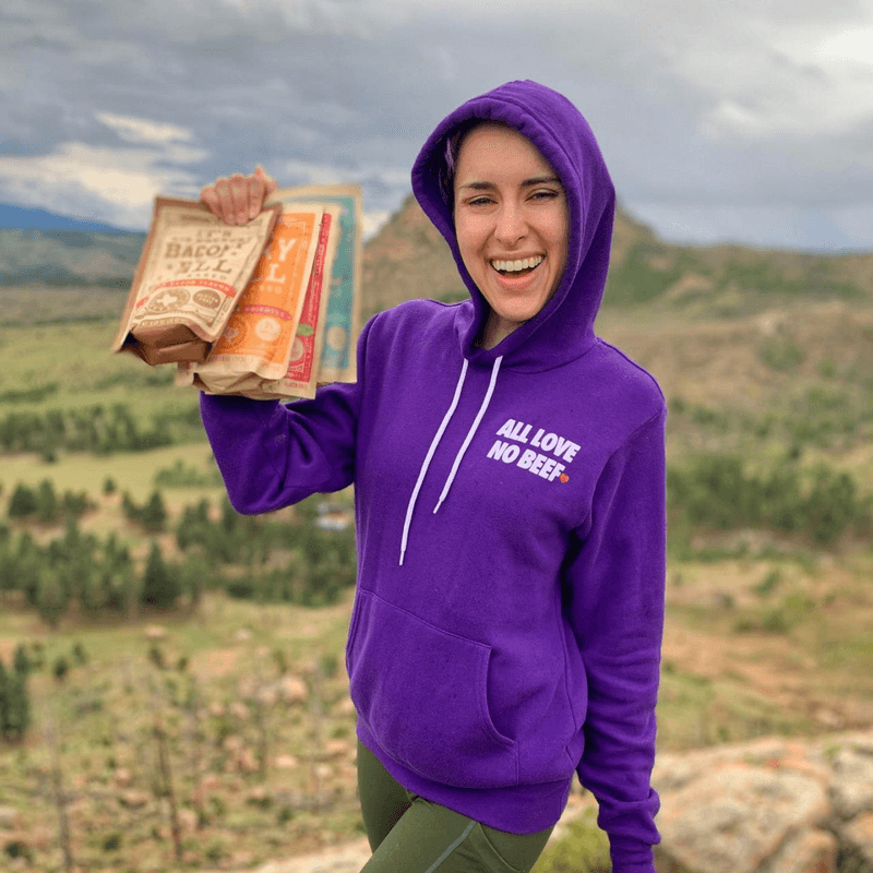 A woman in a purple hoodie that reads 'All Love, No Beef' holding a variety of All Y’alls Foods vegan snacks with a scenic outdoor backdrop.