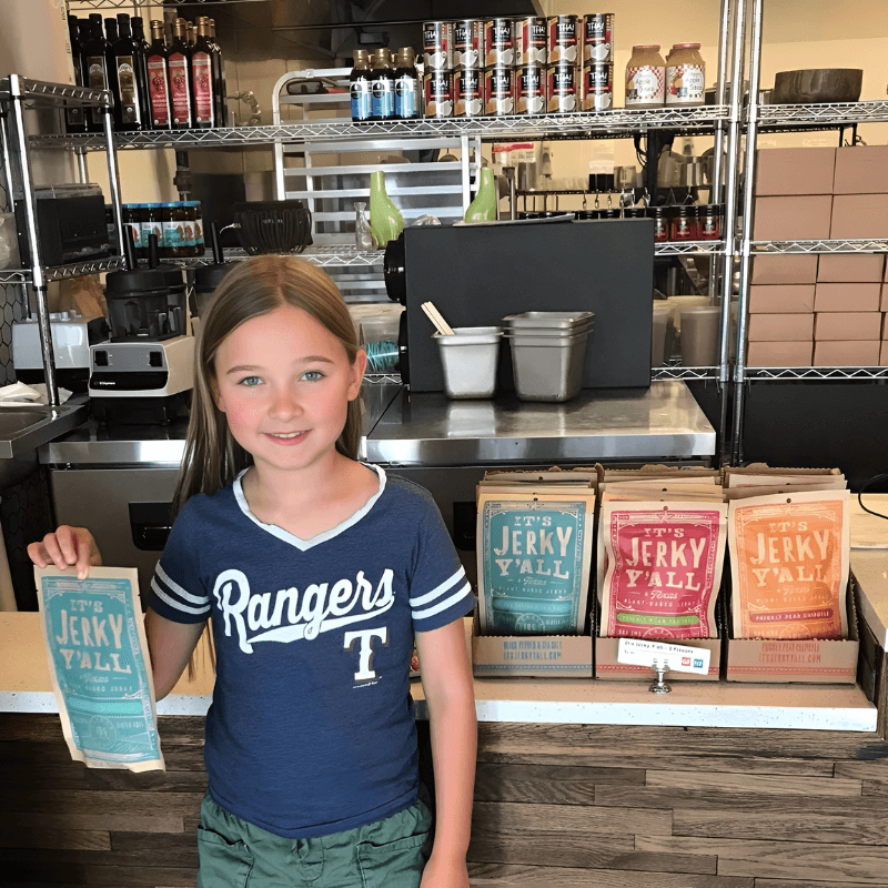  A young girl in a "Rangers" shirt proudly holds a package of "All Y'alls Foods" vegan jerky in a store setting, with various flavors of the plant-based snack displayed beside her