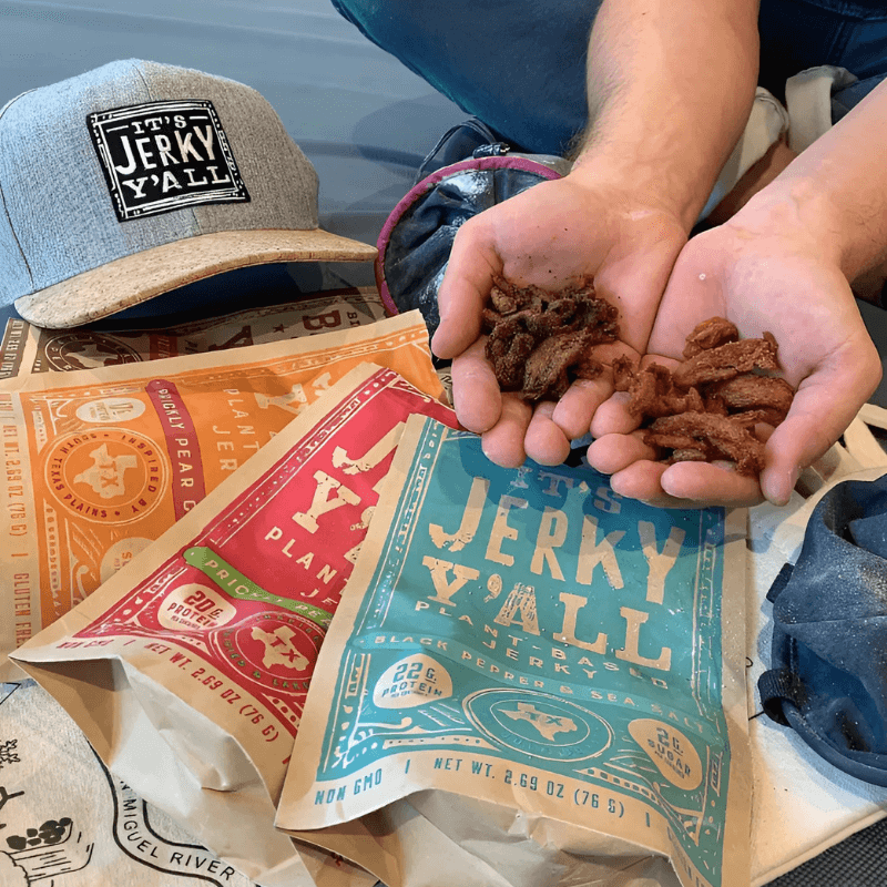 A selection of It's Jerky Y'all plant-based jerky in various flavors, displayed with a branded cap, highlighting high-protein, meat-free snacking options.