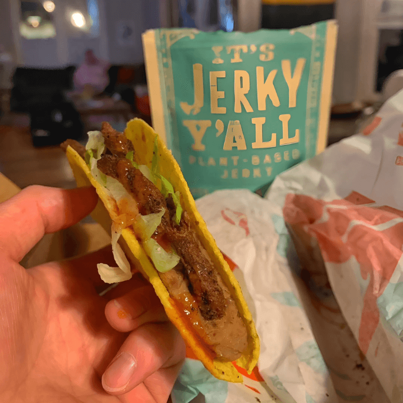 A close-up of a crispy taco filled with fresh lettuce, beans, and plant-based jerky, complemented by packaging in the background promoting a vegan jerky brand.