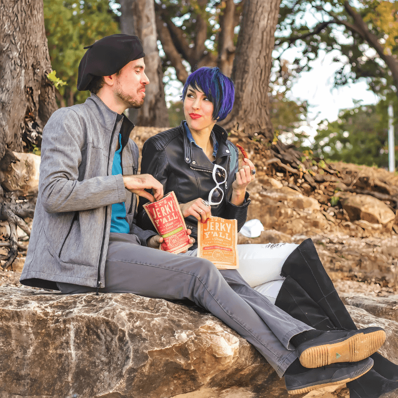 A couple enjoying plant-based jerky outdoors with "It's Jerky Y'all" packs, highlighting the joy of vegan snacks.