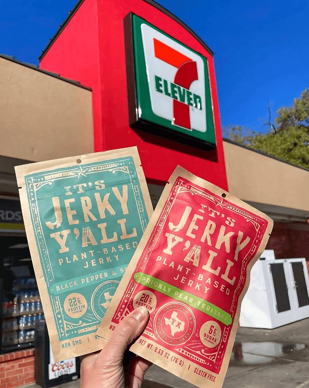 wo packets of plant-based jerky from All Y’alls Foods, displayed in front of a 7-Eleven store, showcasing their availability at major convenience stores.