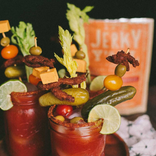 Two Bloody Mary cocktails garnished with skewers of vegan beef jerky, cheese, olives, pickles, and cherry tomatoes, with a bag of 'It's Jerky Y'all' by All Y'all's Foods in the background.