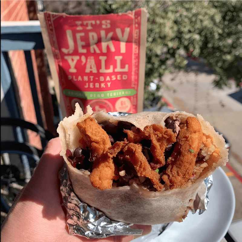 A flavorful burrito filled with vegan jerky, black beans, rice, and fresh ingredients, with a bag of It's Jerky Y'all plant-based jerky in the background, captured outdoors.